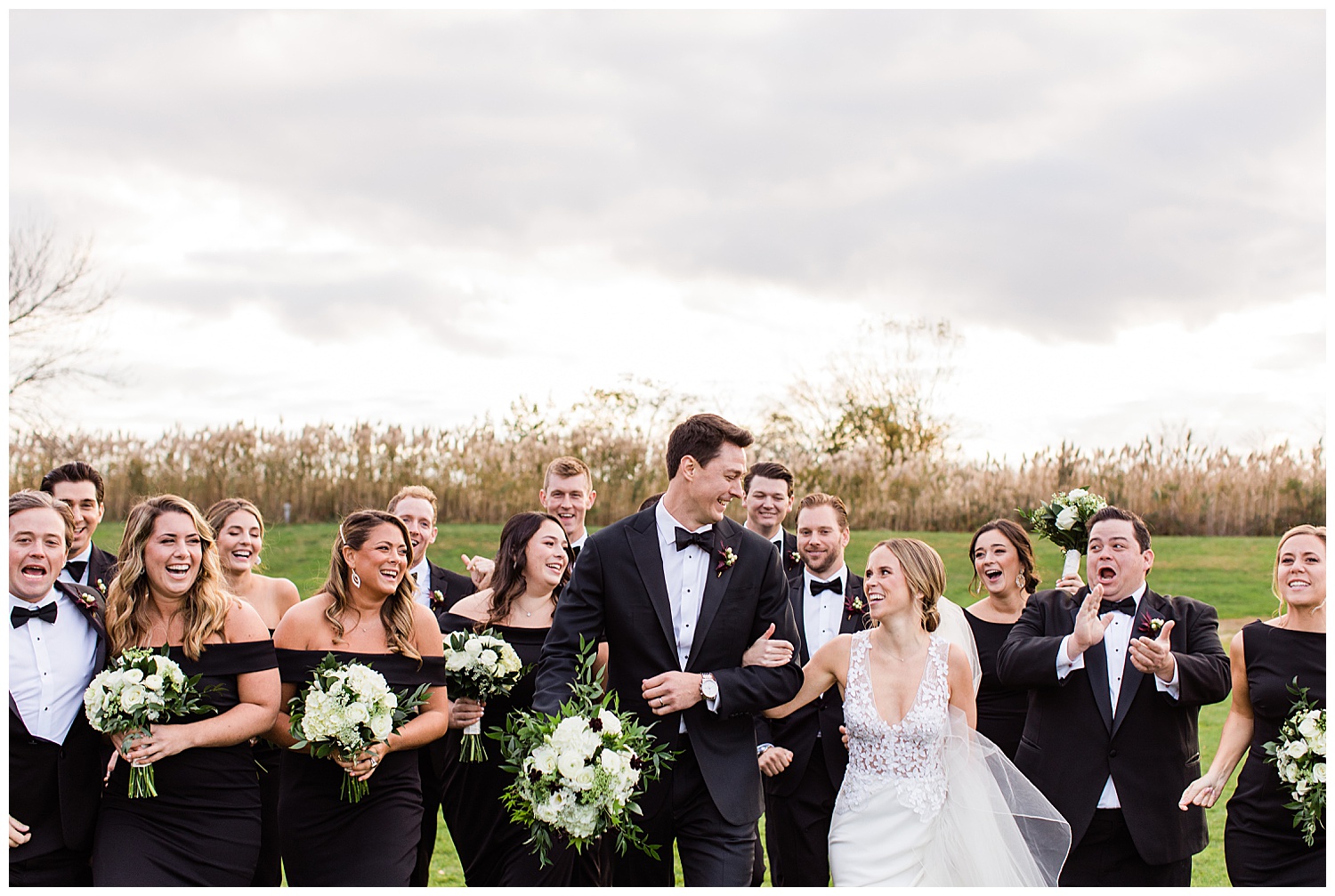 Bride and Groom laughing and smiling at each other as the walk in front of tall grasses with their bridal party at Rumson Country Club Wedding