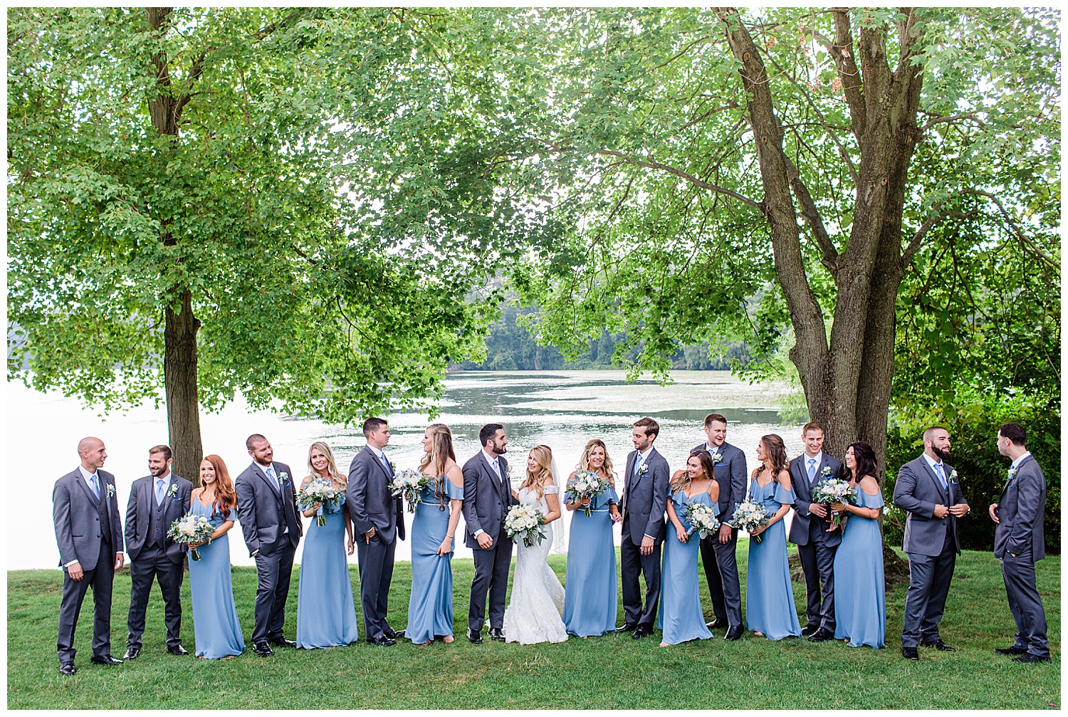 large wedding party laughs in front of lake under big tree