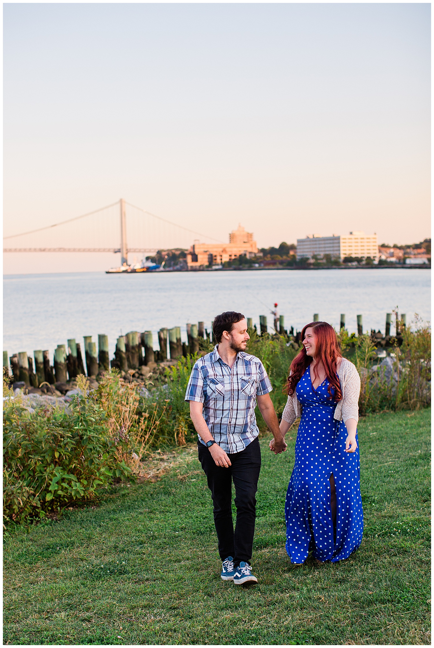 Couple holds hands in field with city bridge in sunset behind them