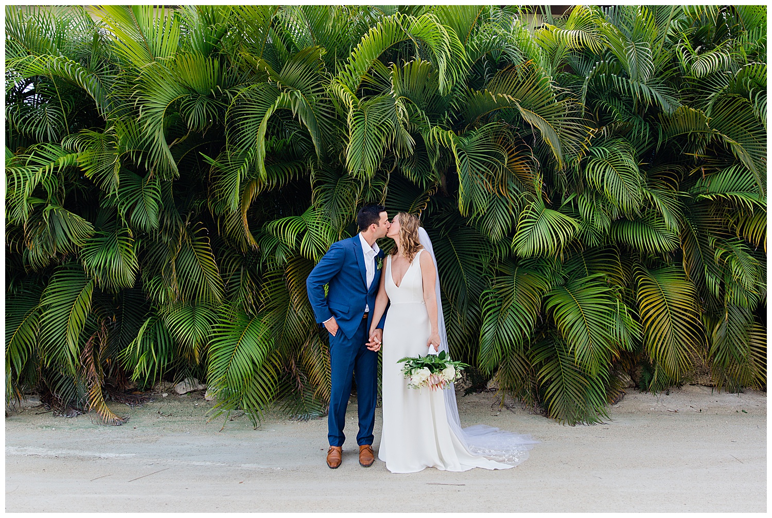 bride and groom kiss in front of wall of green palms