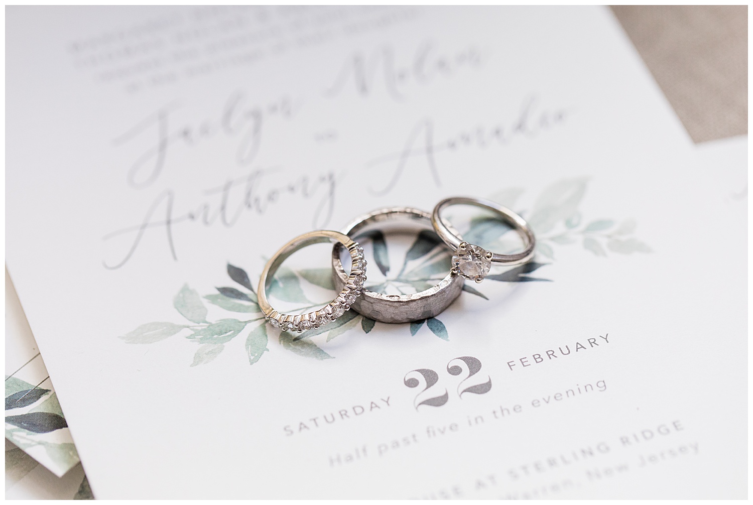 Bride and groom rings stacked on white wedding invitation with greenery